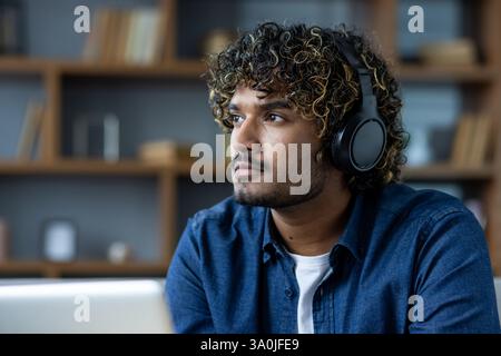 Junge Erwachsene, die Kopfhörer tragen, schauen nachdenklich in die Ferne, tragen ein blaues Jeansshirt vor einem Bücherregal innen. Stockfoto