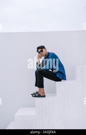 Ein nachdenklicher junger Mann mit Sonnenbrille und blauem Hemd, der draußen auf minimalistischen weißen Stufen sitzt. Er wirkt nachdenklich und entspannt. Der Hintergrund Stockfoto