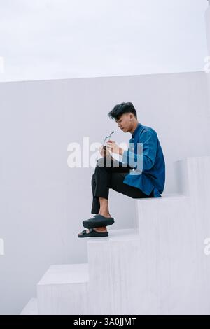 Ein nachdenklicher junger Mann sitzt auf einer minimalistischen weißen Treppe und hält seine Brille. Er trägt lässige, aber stilvolle Kleidung, verkörpert einen modernen, Our Stockfoto