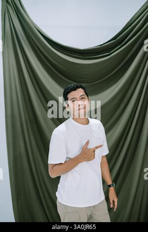 Ein asiatischer Mann in einem weißen T-Shirt zeigt mit dem Finger, präsentiert oder schlägt etwas vor. Er steht vor dem grünen Vorhang. Verwenden Sie diese Stockfoto