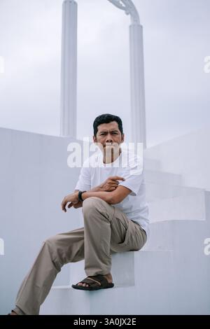 Ein Mann sitzt nachdenklich auf weißen Stufen in der Nähe von monumentalen Säulen in einer urbanen Umgebung. Er trägt ein schlichtes Outfit, sein Ausdruck deutet auf Kontemplat hin Stockfoto