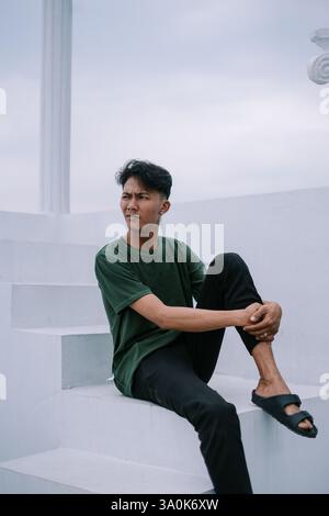 Ein junger Mann sitzt nachdenklich auf weißen minimalistischen Stufen draußen. Er ist lässig in dunkler Kleidung gekleidet und erscheint tief in Gedanken und blickt in die Th Stockfoto