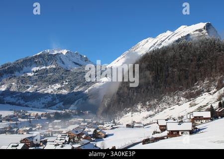 Winteralpinpanorama in Au, Österreich - schneebedeckte Berge, nebelige Wälder und ein idyllisches Bergdorf in der Sonne. Stockfoto