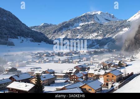 Malerisches Alpendorf im Winter – schneebedeckte Chalets, majestätische Berge, blauer Himmel und neblige Täler – perfektes Winterwunderland. Stockfoto