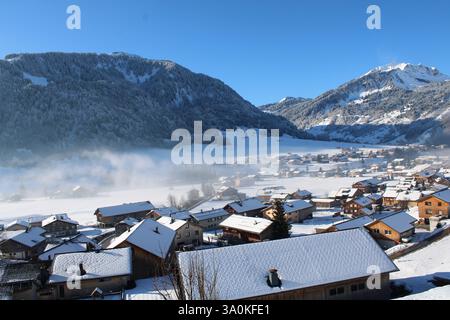 Winteralpinpanorama in Au, Österreich - schneebedeckte Berge, nebelige Wälder und ein idyllisches Bergdorf in der Sonne. Stockfoto