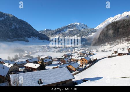 Winterliches Alpenpanorama in Au, Österreich – Schneebedeckte Berge, nebelverhangene Wälder und ein idyllisches Bergdorf im Sonnenschein. Stockfoto