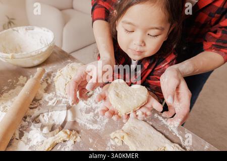 Mutter und Kind backen zusammen, Hände halten herzförmigen Teig, Familienbindung, hausgemachtes Gebäck, gemütliche Küchenszene, mehlbedeckte Hände. Stockfoto