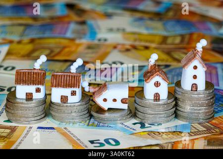 Fünf Miniaturhäuser auf einem Stapel von Münzen unterschiedlicher Höhe, ein Haus ist auf die Seite gefallen Stockfoto