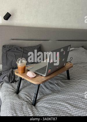 Ein Laptop mit verschiedenen Aufklebern befindet sich auf einem klappbaren Holztisch mit schwarzen Beinen. Eine pinkfarbene kabellose Maus und ein Eiskaffee in einer Plastiktasse Stockfoto