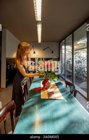 Eine Frau in einem minimalistischen Kleid dekoriert einen rustikalen Esstisch mit Blumenarrangements in einer modernen Küche. Große Glasfenster sorgen für sanftes natürliches Licht und unterstreichen die warme und ruhige Atmosphäre Stockfoto