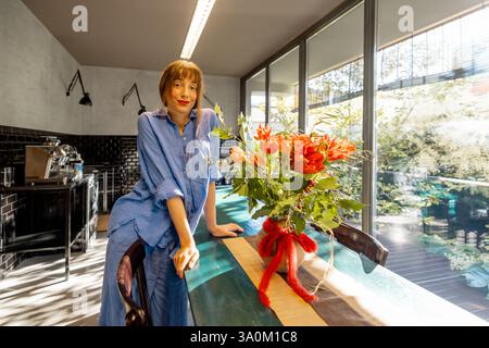 Eine Frau in einem entspannten blauen Hemd ziert einen rustikalen Esstisch mit Blumenarrangements in einer stilvollen Küche. Tageslicht durch große Glasfenster verstärkt die warme und einladende Atmosphäre Stockfoto