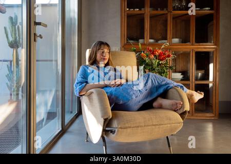 Eine Frau in blauem Leinen-Outfit entspannt sich in einem Vintage-Sessel neben einem sonnendurchfluteten Fenster. Das moderne Interieur, der Holzschrank und die frischen Blumen schaffen eine warme, gemütliche und inspirierende Atmosphäre des langsamen Lebens Stockfoto