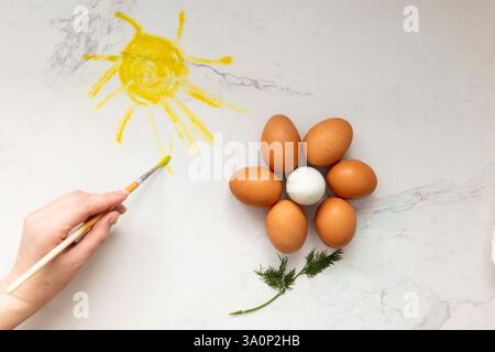 Eine geschickte Hand malt fachmännisch die strahlende Sonne mit wunderschönen bunten Eiern, die eine helle Blume auf glattem Marmor bilden, eine Osterdekoration Stockfoto
