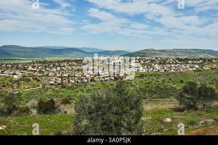 Ein Panoramablick auf eine ländliche Stadt, eingebettet zwischen sanften grünen Hügeln, unter einem hellblauen Himmel, die Natur und menschliche Siedlung zeigt Stockfoto