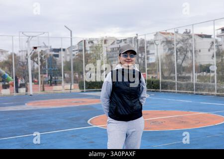Eine Frau mittleren Alters steht selbstbewusst auf einem Basketballfeld und trägt sportliche Freizeitkleidung. Der Park im Hintergrund bietet farbenfrohe Spielplätze Stockfoto
