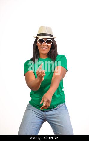 Die stilvolle schwarze Frau tanzt und zeigt mit dem Finger, während sie ein grünes T-Shirt, eine Sonnenbrille und einen Hut vor weißem Hintergrund trägt Stockfoto