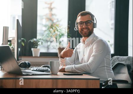 Lächelnd, am Tisch sitzend. In Kopfhörern. Büroangestellte in formeller Kleidung sitzt am Computer und arbeitet. Stockfoto