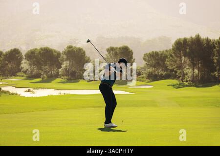 Ein Golfspieler, der geschickt einen Club schwingt und an einem hellen, sonnigen Tag den Ball in der Nähe des Sandbunkers anvisiert. Die üppige grüne Umgebung unterstreicht den ruhigen Golfplatz Stockfoto