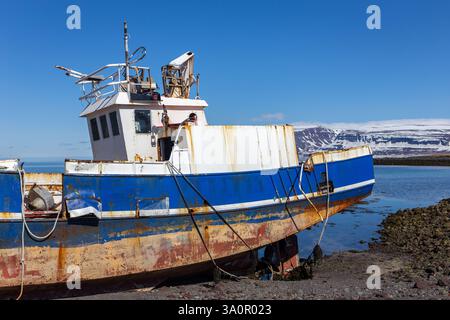Altes, rostiges Stahlfischboot, das am Ufer der Westfjorde, Island, mit schneebedeckten Bergen im Hintergrund liegt. Stockfoto