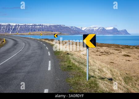 Gewundene Asphaltstraße durch die Westfjorde in Island mit schneebedeckten Bergen und Meerblick im Hintergrund. Stockfoto