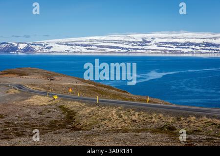 Gewundene Asphaltstraße durch die Westfjorde in Island mit schneebedeckten Bergen und Meerblick im Hintergrund. Stockfoto