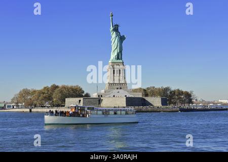 Die Freiheitsstatue auf der Insel mit einem klaren blauen Himmel im Hintergrund an einem sonnigen Tag, Manhattan, Downtown, New York City, Touristenboot auf dem Wasser Stockfoto