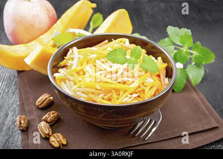 Kürbis-, Karotten- und Apfelsalat mit Pekannüssen, gewürzt mit Pflanzenöl in einer Schüssel auf einem Handtuch, Minze auf dunklem Holzbrett-Hintergrund Stockfoto