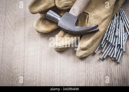 Schutzhandschuhe aus Leder mit Klauenhammer und Haufen von Metallnägeln auf Holzbrett in Nahaufnahme Konstruktionskonzept Stockfoto