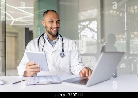 Junger erfahrener Arzt, der in der Klinik mit Tablet-Computer und Laptop arbeitet. Medic in weißem medizinischem Mantel und mit Stethoskop, das Patienten aus der Ferne hilft. Stockfoto