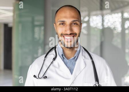 Zufriedener erfolgreicher Arzt in Nahaufnahme lächelnd und in die Kamera schauend. Arzt in weißem medizinischem Mantel und mit Stethoskop im Büro am Arbeitsplatz, moderne Klinik. Stockfoto