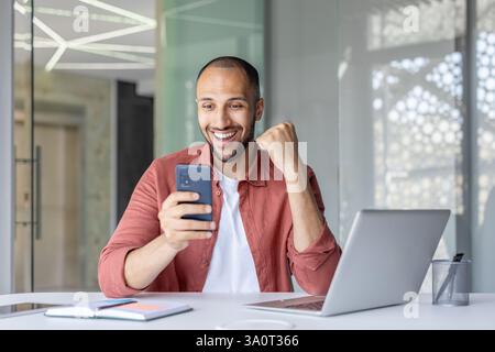 Erfolgreicher Mann mit Telefon in den Händen, der Sieg und triumphale Erfolge feiert. Geschäftsmann erhielt eine Benachrichtigung auf dem Smartphone, saß mit Laptop im Büro. Stockfoto