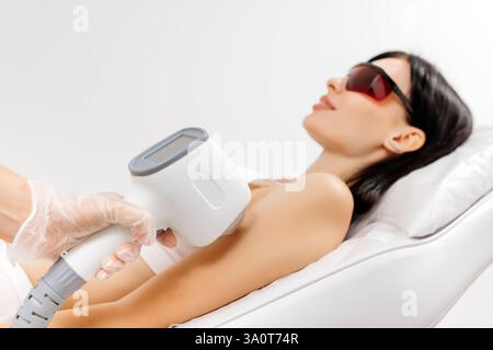 Körperpflegekonzept. Laserhaarentfernung. Junge weiße Frau in Schutzbrille liegt auf dem Sofa, weibliche Kosmetikerin, die Laser-Haarentfernung macht Stockfoto