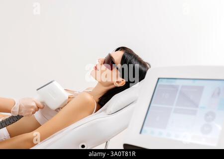 Körperpflegekonzept. Laserhaarentfernung. Junge weiße Frau in Schutzbrille liegt auf dem Sofa, weibliche Kosmetikerin, die Laser-Haarentfernung macht Stockfoto