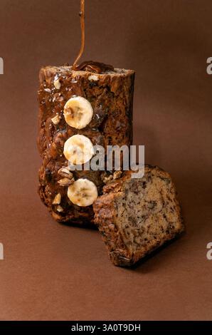 Modernes Stillleben mit hausgemachtem Bananenbrot. Brot mit Walnüssen, mit Karamell getränkt, aufrecht stehend auf braunem Hintergrund. Soße von oben ausgießen Stockfoto