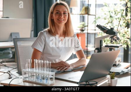 Lächelnde, fröhliche Stimmung. Junge Ärztin im Mantel sitzt in der Klinik und arbeitet. Stockfoto