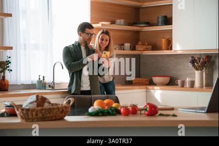 Mann, der der Frau am Telefon Medien zeigt. Das junge Paar ist zusammen in der Küche. Stockfoto
