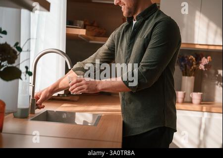 Gießt Wasser aus dem Wasserhahn in ein Glas. Ein gutaussehender Mann ist tagsüber in der Küche. Stockfoto