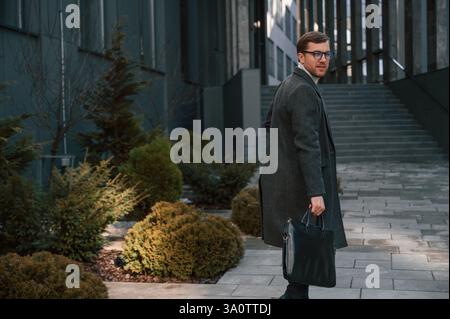 Schauend und spazierend. Hübscher Mann in formeller Kleidung ist draußen in der Nähe des Geschäftsgebäudes. Stockfoto