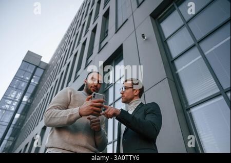 Smartphone halten. Geschäftsmann in formeller Kleidung ist draußen mit schwarzem Mann in der Stadt. Stockfoto