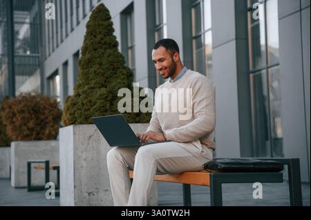 Mit Laptop auf der Bank sitzen. Ein schöner schwarzer Mann ist draußen in der Nähe des Geschäftsgebäudes. Stockfoto
