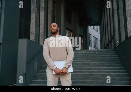 Vorderansicht. Mit dem Laptop in der Hand stehen. Ein schöner schwarzer Mann ist draußen in der Nähe des Geschäftsgebäudes. Stockfoto