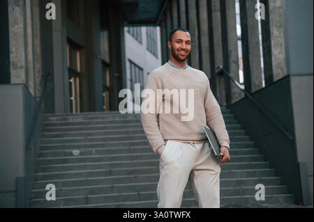Lächelnd und hält den Laptop in der Hand. Ein schöner schwarzer Mann ist draußen in der Nähe des Geschäftsgebäudes. Stockfoto