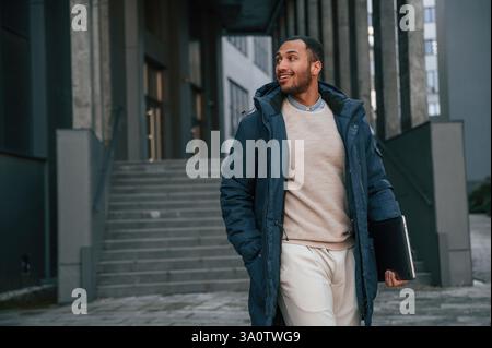 In blauer Jacke und mit Laptop in den Händen. Ein schöner schwarzer Mann ist draußen in der Nähe des Geschäftsgebäudes. Stockfoto