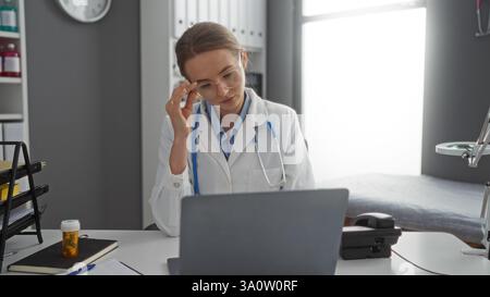 Frau Ärztin sitzt in der Klinik mit einer Brille, die nachdenklich auf den Laptop blickt umgeben von medizinischen Geräten und Dokumenten mit Uhr an der Wand in hell Stockfoto