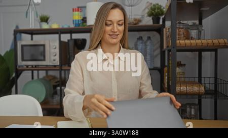 Junge Frau mit blonden Haaren, die in einem Büro arbeitet und Dokumente an ihrem Schreibtisch arrangiert, umgeben von moderner Einrichtung und Regalen mit verschiedenen IT-Elementen Stockfoto