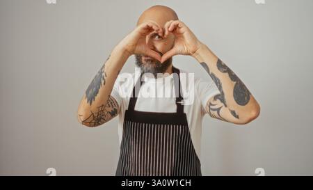 Hübscher hispanischer Mann mit Bart und Tätowierungen, mit gestreifter Schürze, mit den Händen über einem isolierten weißen Hintergrund eine Herzform Stockfoto
