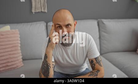 Hispanischer Mann mit Bart und Tätowierungen, der in einem Wohnzimmer sitzt, nachdenklich aussieht und ein weißes T-Shirt trägt. Stockfoto