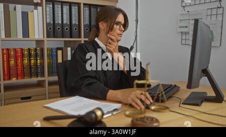 Frau, die an ihrem Schreibtisch in einem Gerichtssaal arbeitet, konzentriert sich auf den Computer, umgeben von Rechtsbüchern und Justizwaagen, mit einem durchdachten E Stockfoto
