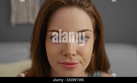 Eine junge Frau mit roten Haaren und blauen Augen sieht zu Hause im Wohnzimmer in die Kamera und zeigt ein attraktives und schönes Gesicht. Stockfoto