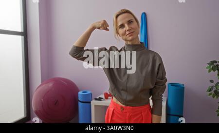 Blonde Frau im Fitnessstudio, die ihren Arm beugt und Stärke und Selbstvertrauen unter den Trainingsgeräten und Fitnessgeräten in einem hellen Innenraum betont Stockfoto